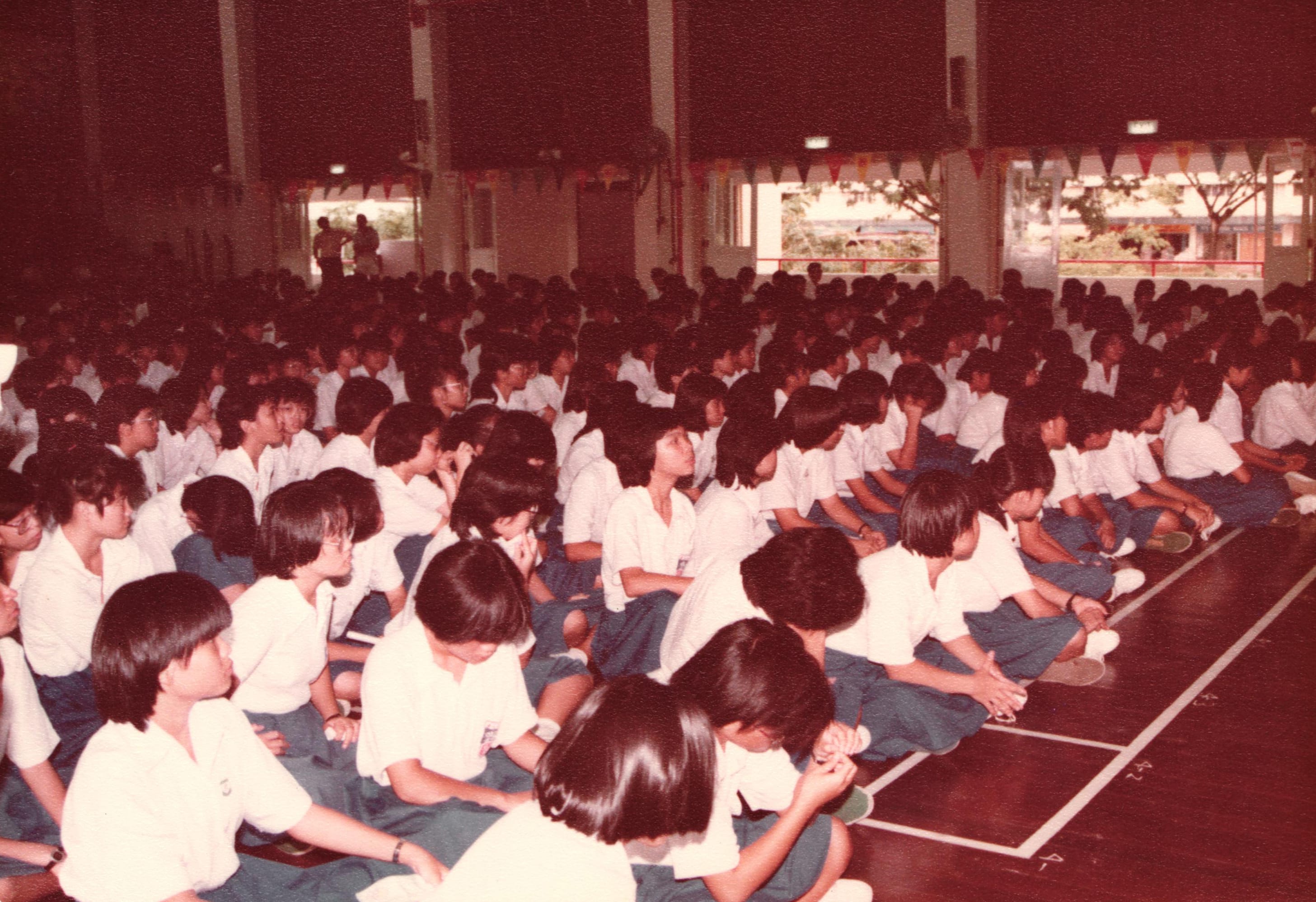 Large group of students in white shirts and blue skirts sitting on a gymnasium floor during a school assembly.
